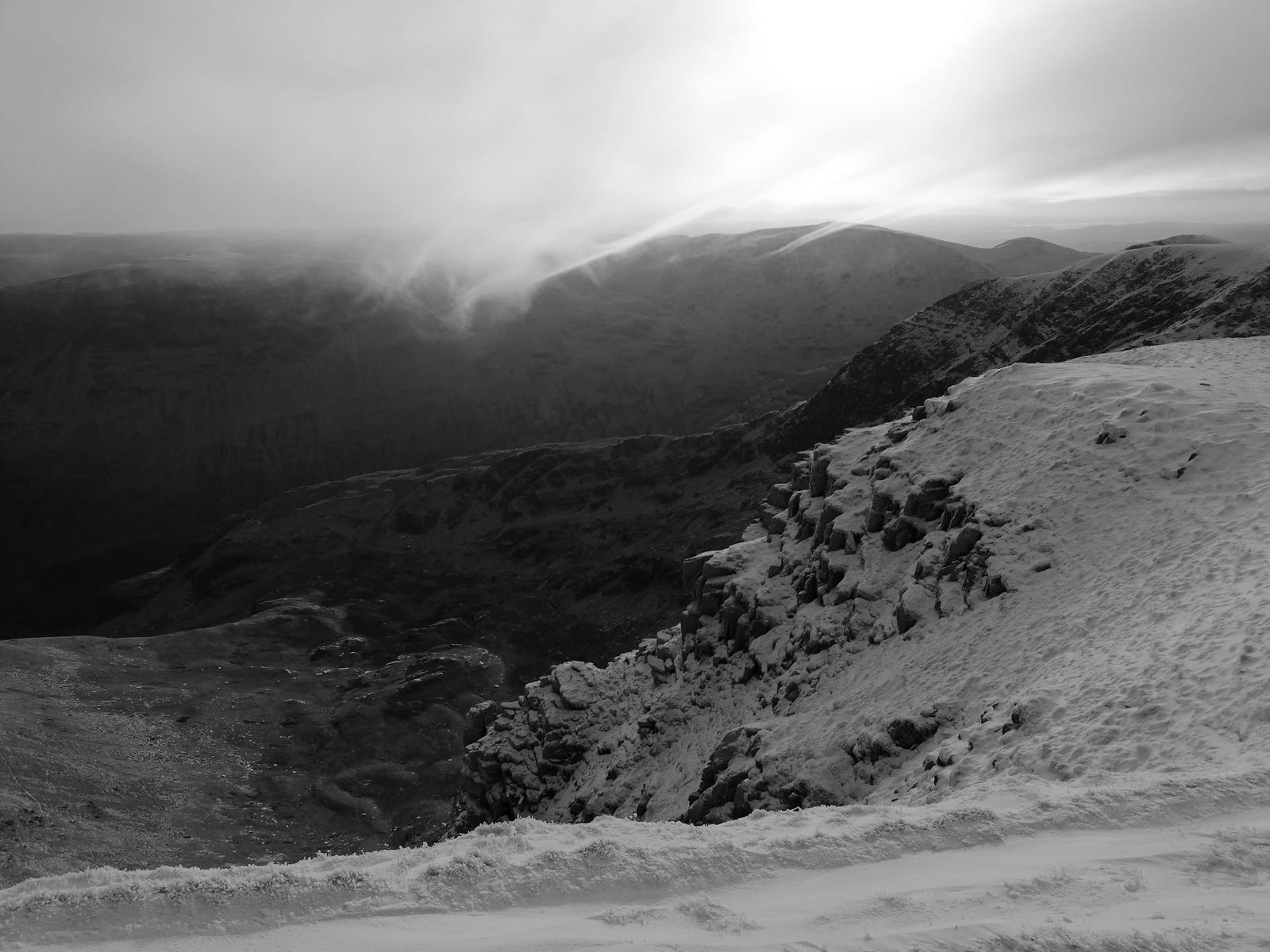Black and white winter view of wind-blown snow along the edge of Helvellyn's high ground, with crags and fells beyond under bright cloud