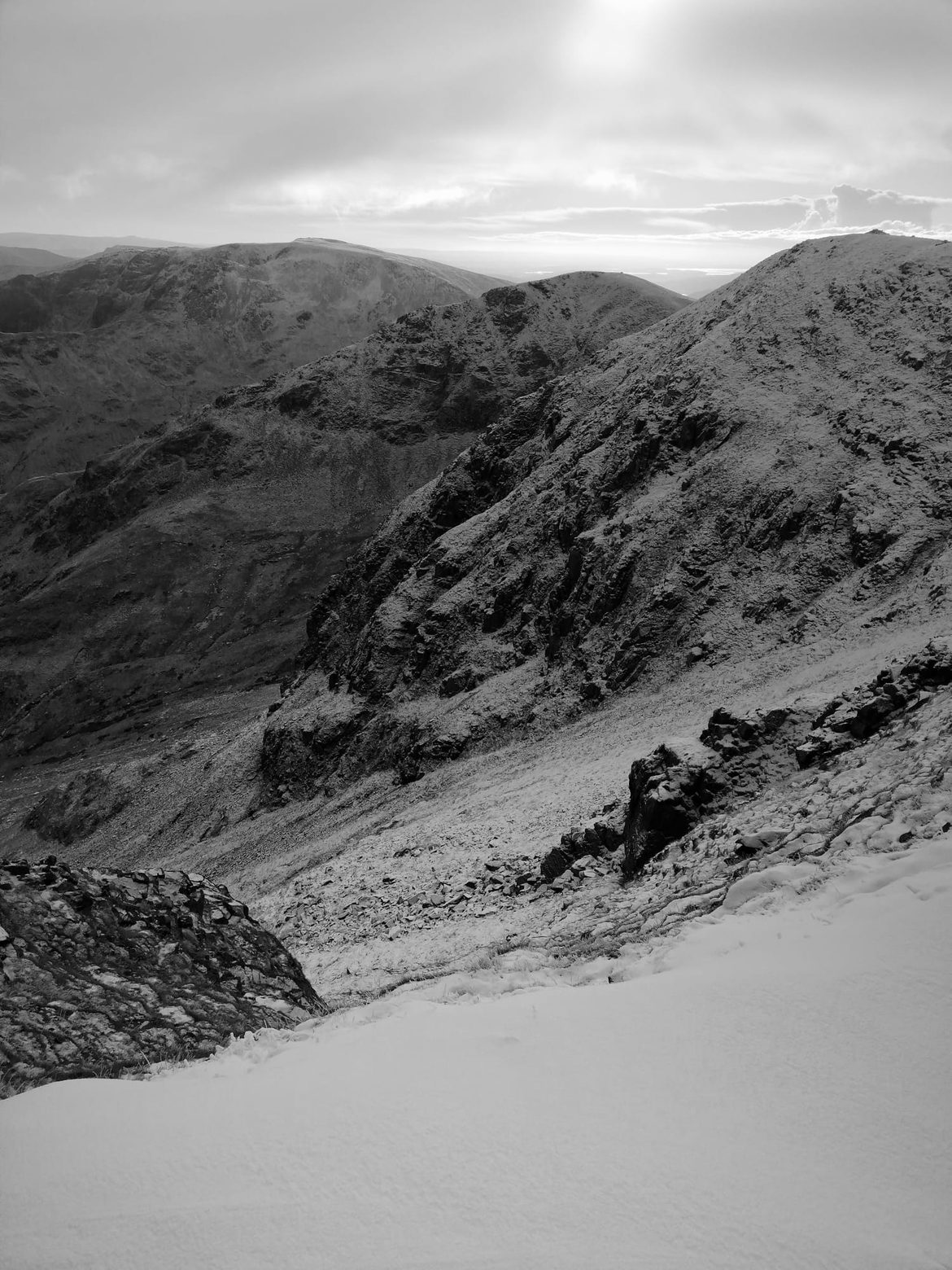 Black and white view across snowy crags and a deep valley from high on Helvellyn, with deep snow in the foreground