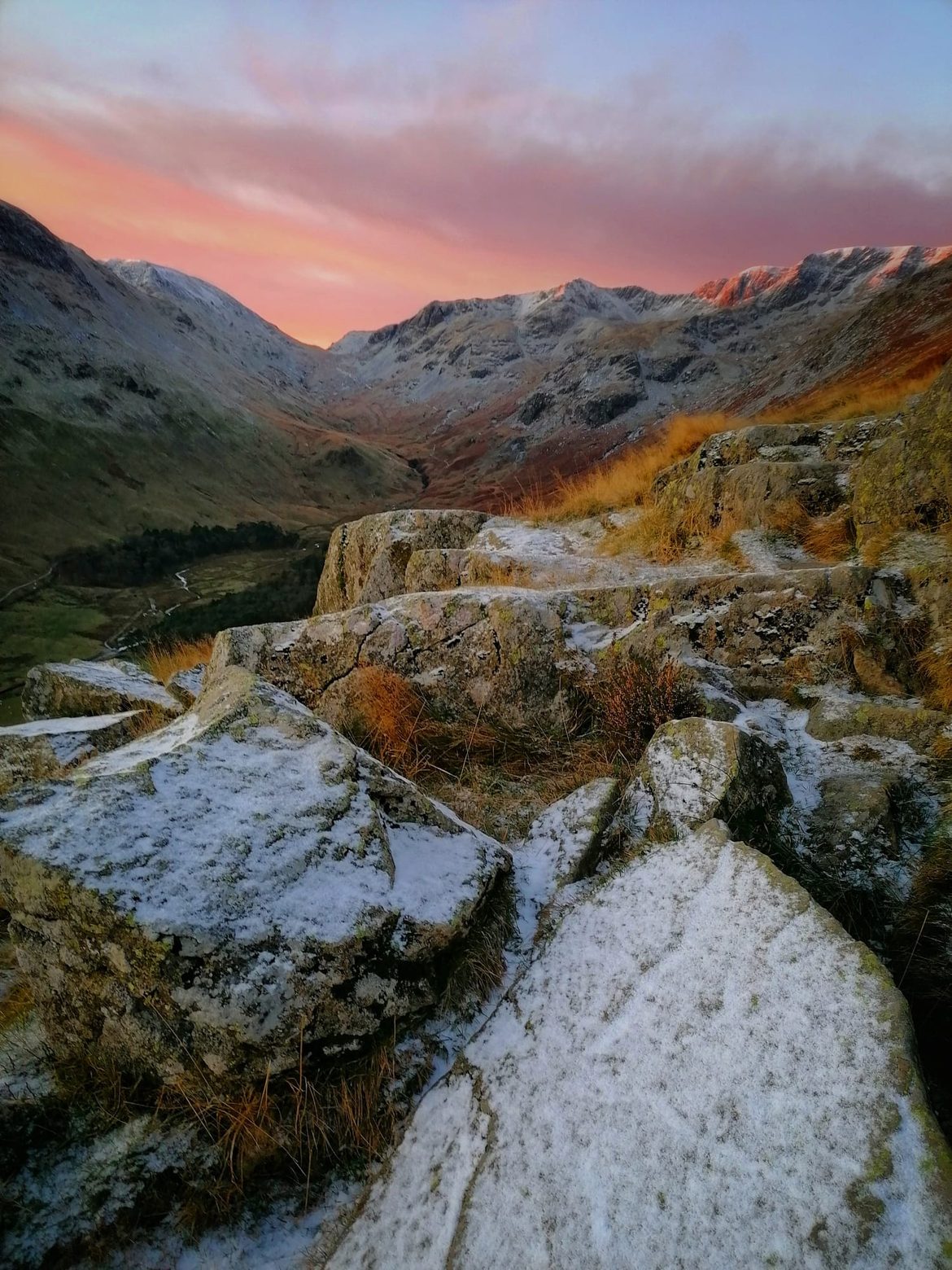 Snow-dusted boulders and golden grass in the foreground, a valley and snowy crags beyond under a pink winter sky, Helvellyn