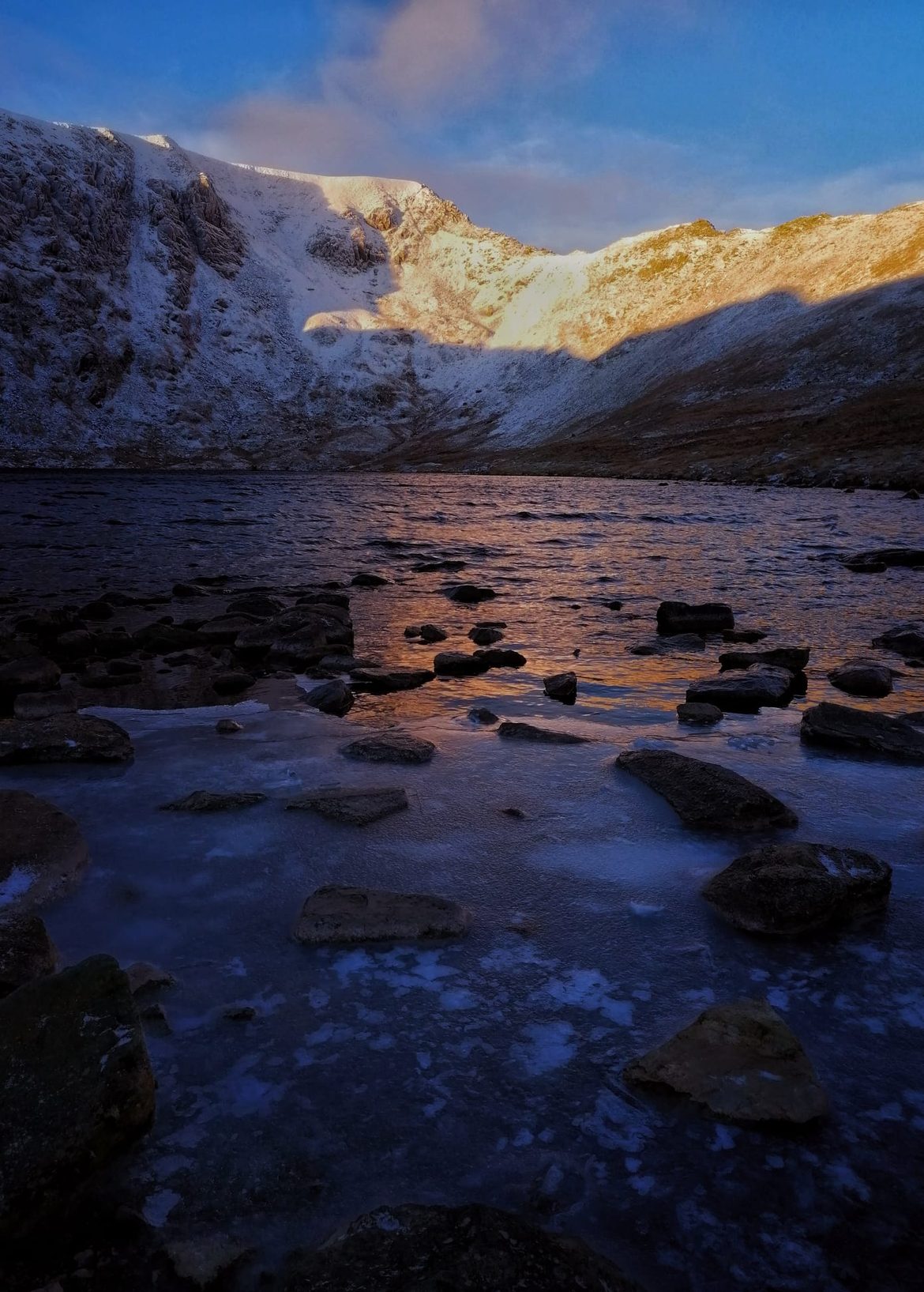 Ice and rocks at the edge of a frozen tarn below Helvellyn, the snowy headwall above catching low sun under a blue sky