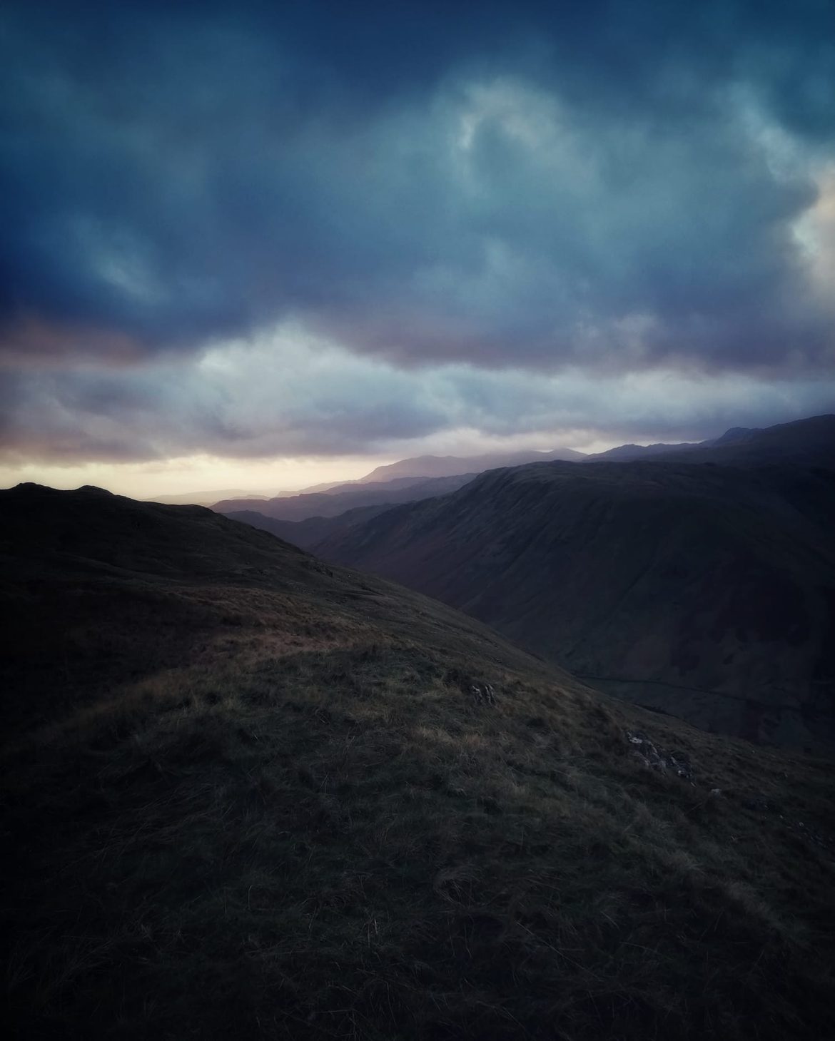 Dark fell ridges above Honister Pass under heavy blue cloud, with a band of evening light on the horizon