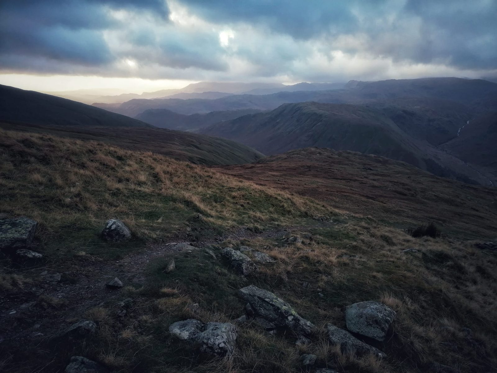 Rough grass and scattered boulders on the fell top above Honister Pass, with layers of fells fading into the distance at dusk
