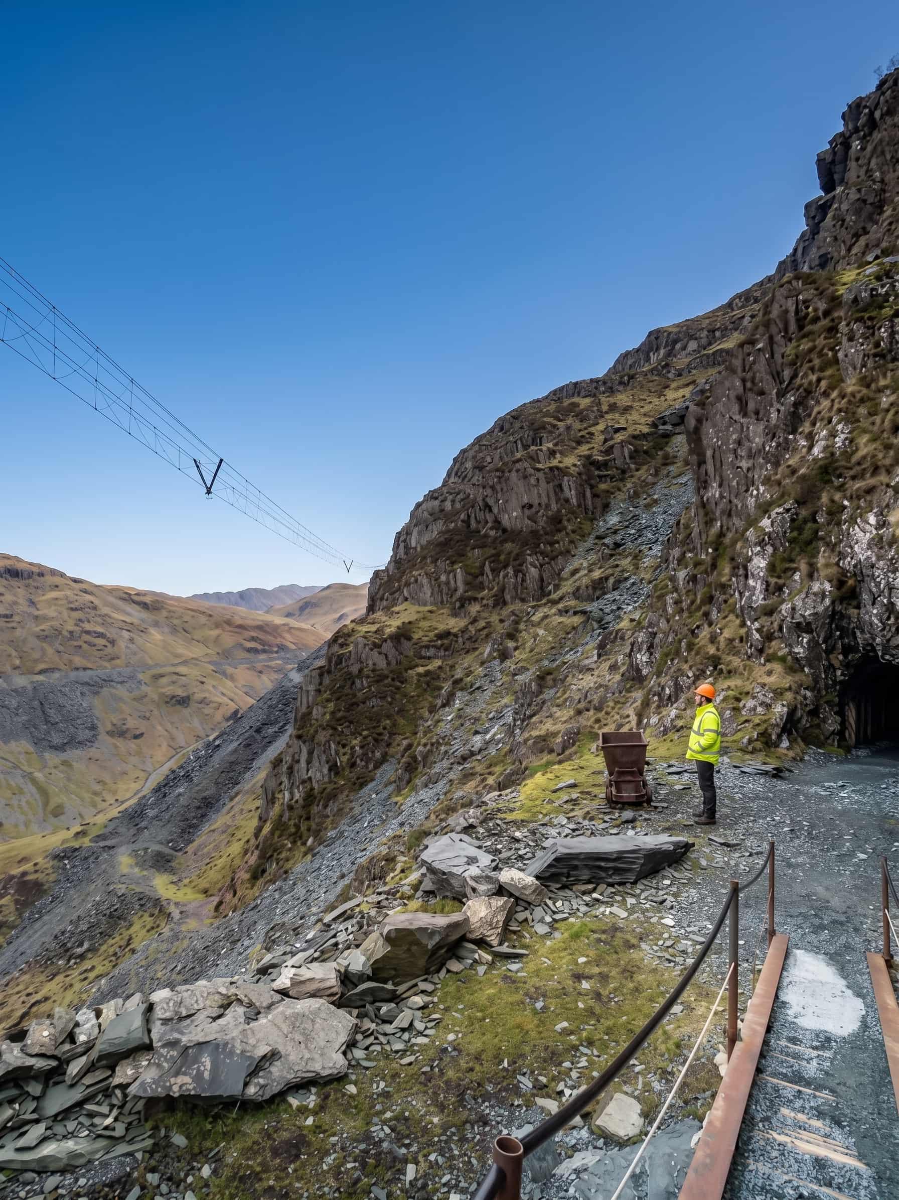 The Aerial Flight cables crossing high above the old mine workings at Honister, with a rusted tipper wagon and a mine entrance on the level