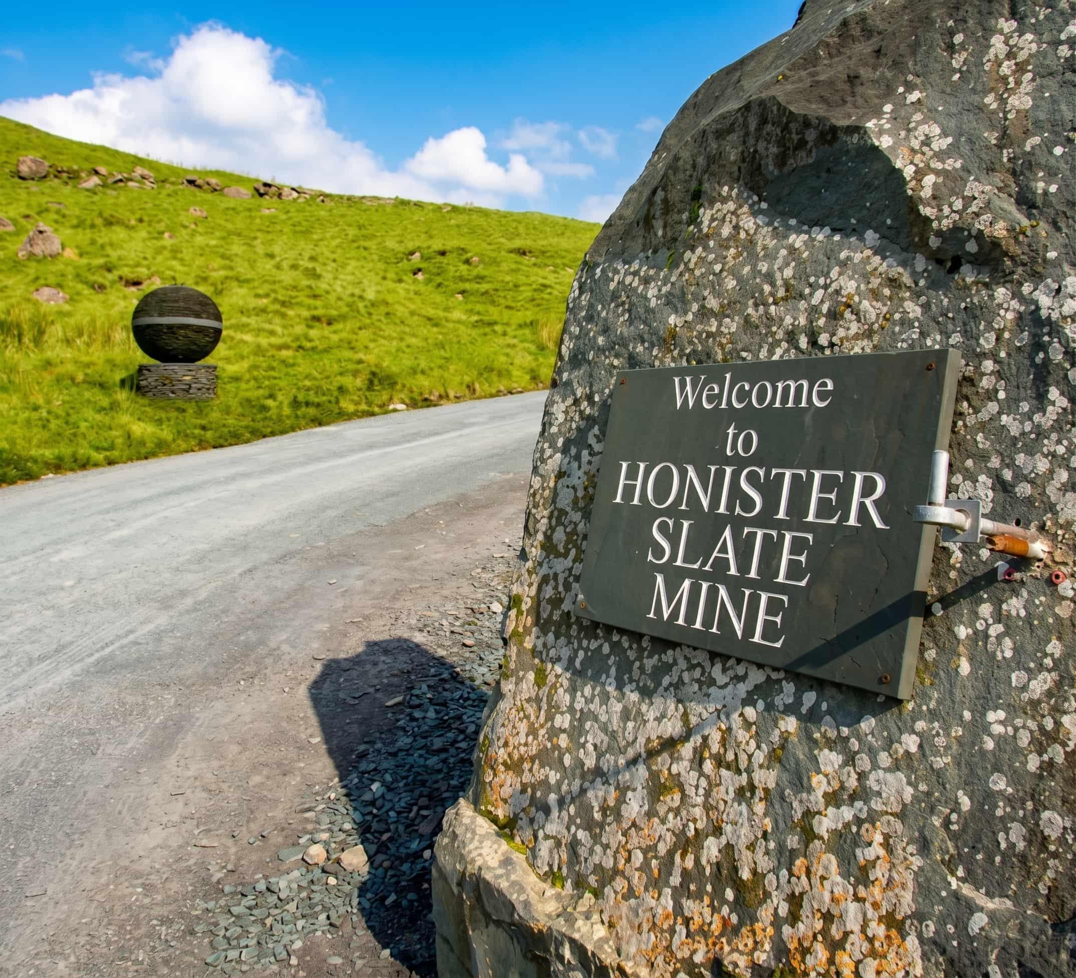 The Welcome to Honister Slate Mine plaque on a boulder at the entrance, with the slate sphere sculpture on the fellside behind