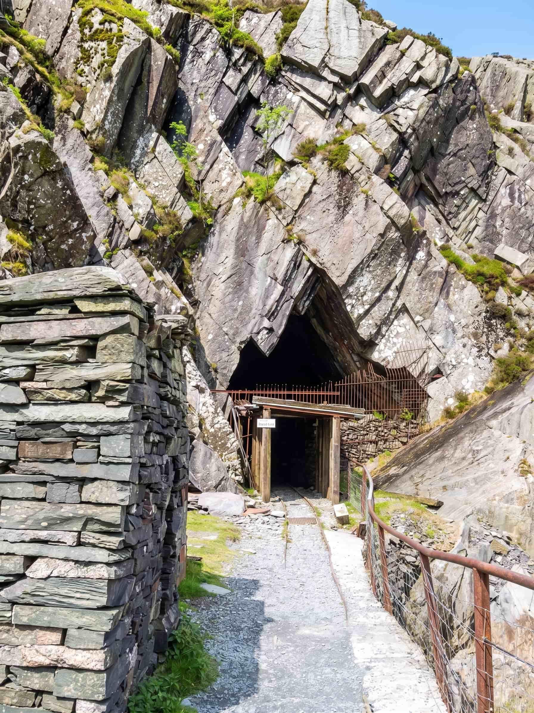 A mine entrance framed by folded slate crags at Honister, with narrow-gauge rails running in past a dry stone slate wall