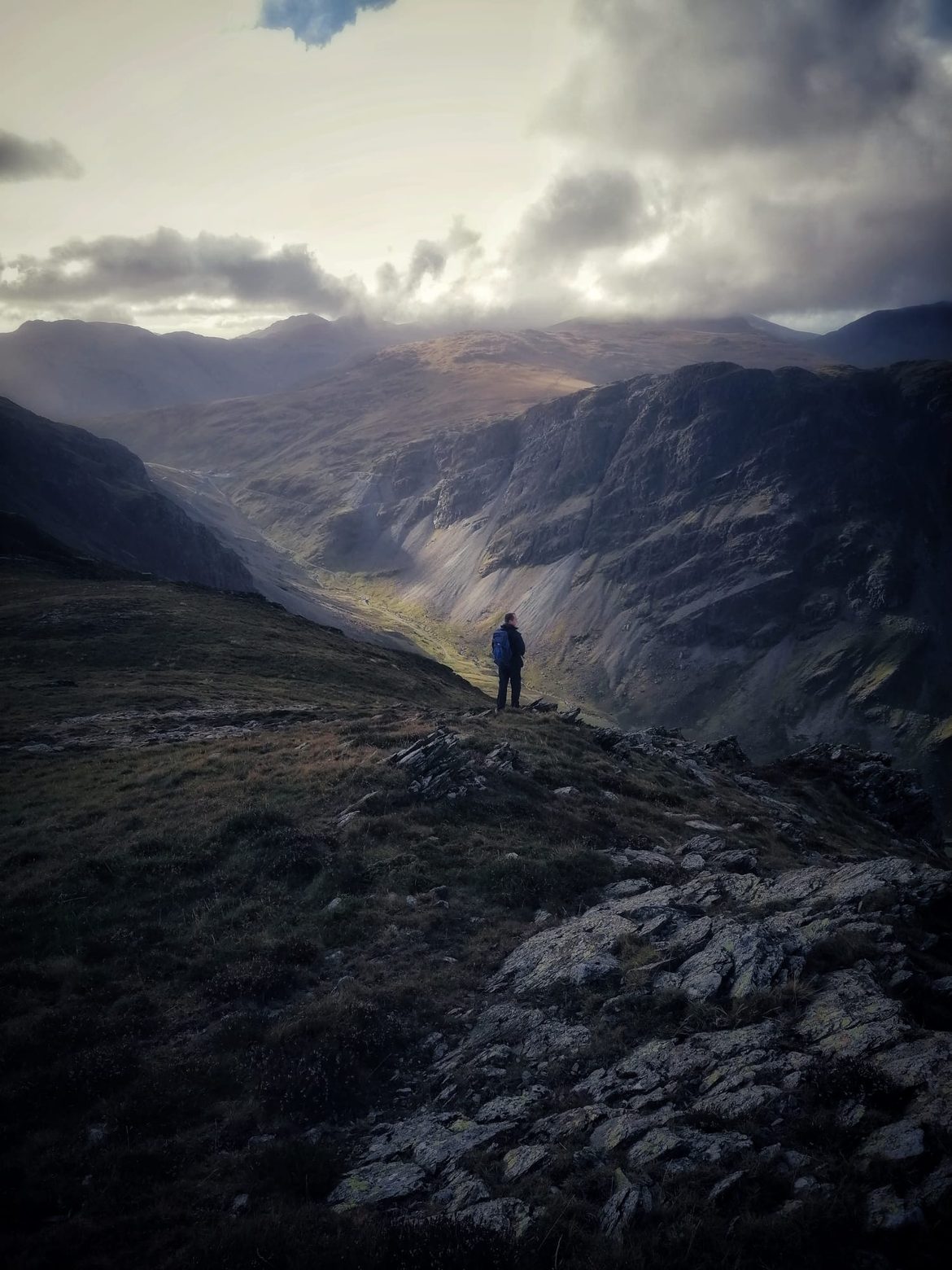 A lone walker standing on a rocky fellside above Honister Pass, looking down a steep-sided valley under a stormy sky