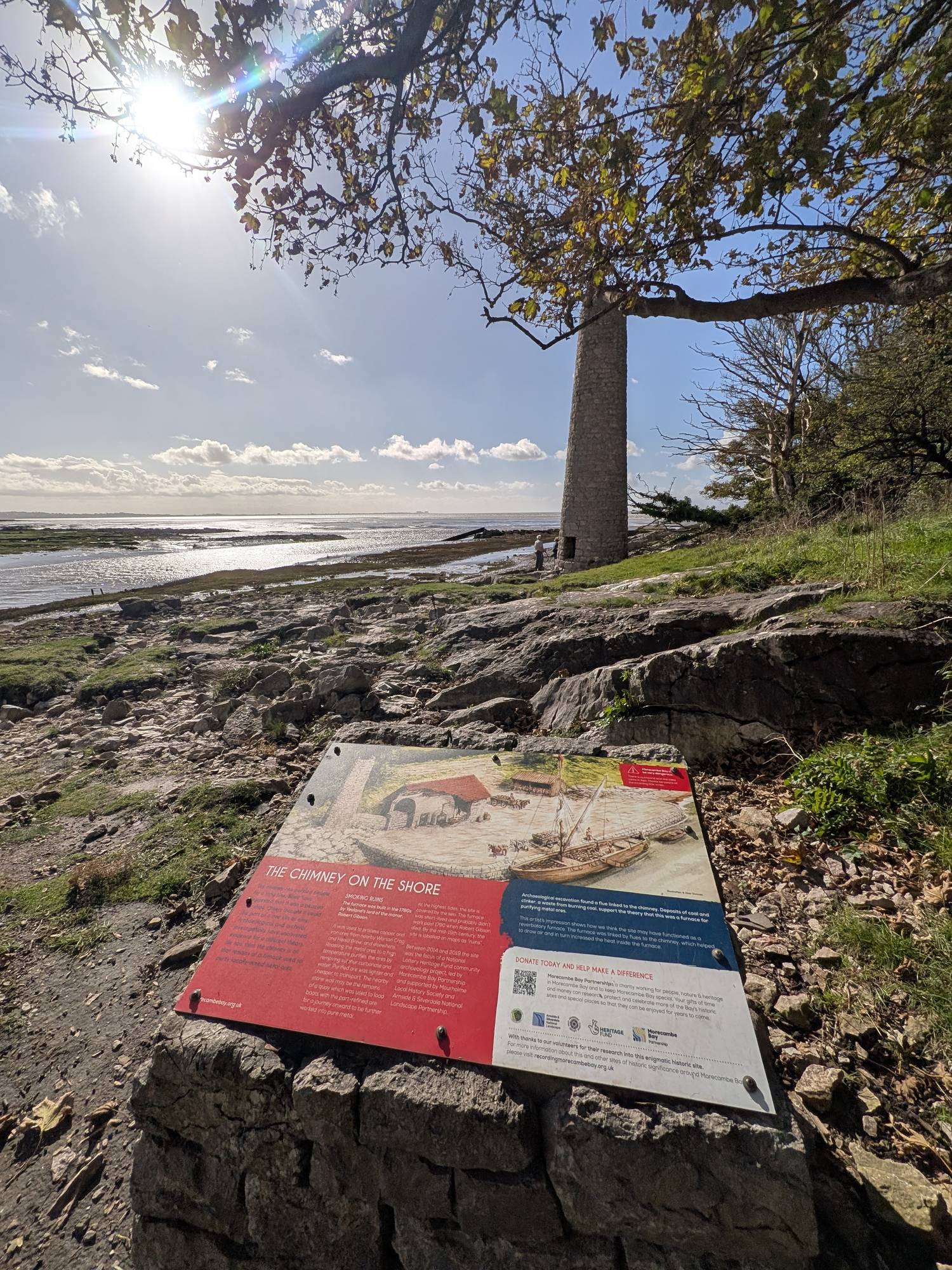 An information board titled The Chimney on the Shore mounted on a stone plinth, with the chimney and Morecambe Bay behind