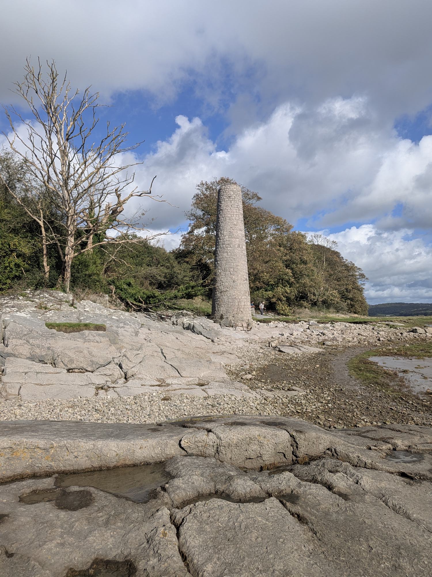 A tall, tapering stone chimney standing on the limestone shore at Jenny Brown's Point, backed by trees