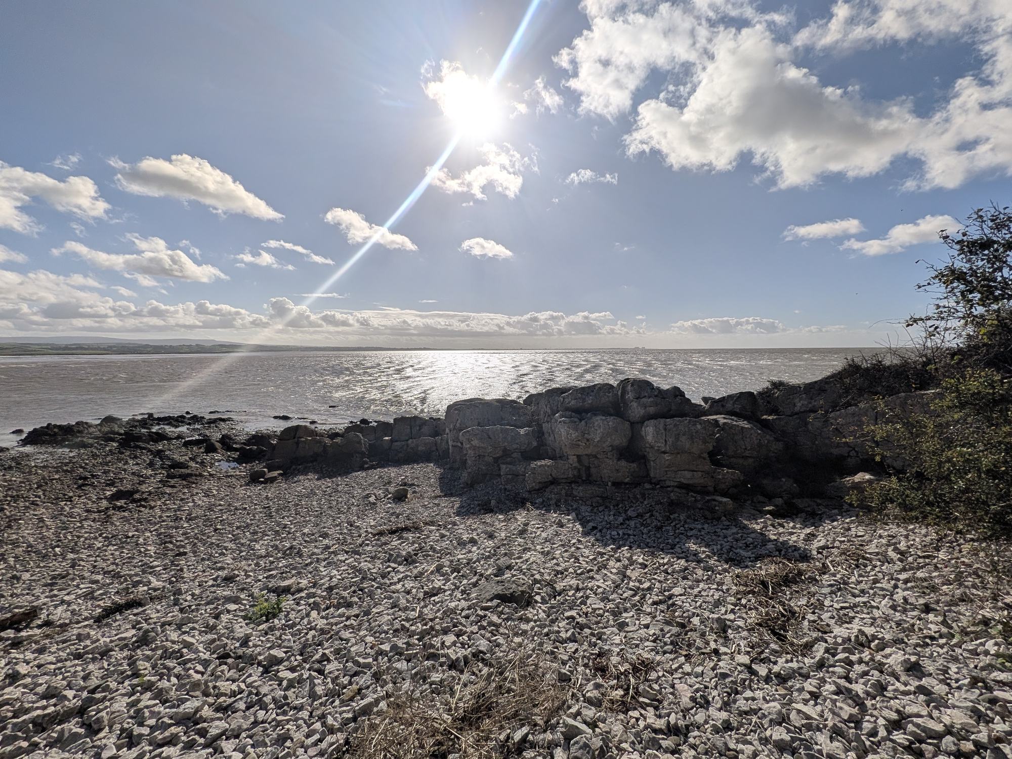 A limestone outcrop on a shingle shore with Morecambe Bay and the sun behind