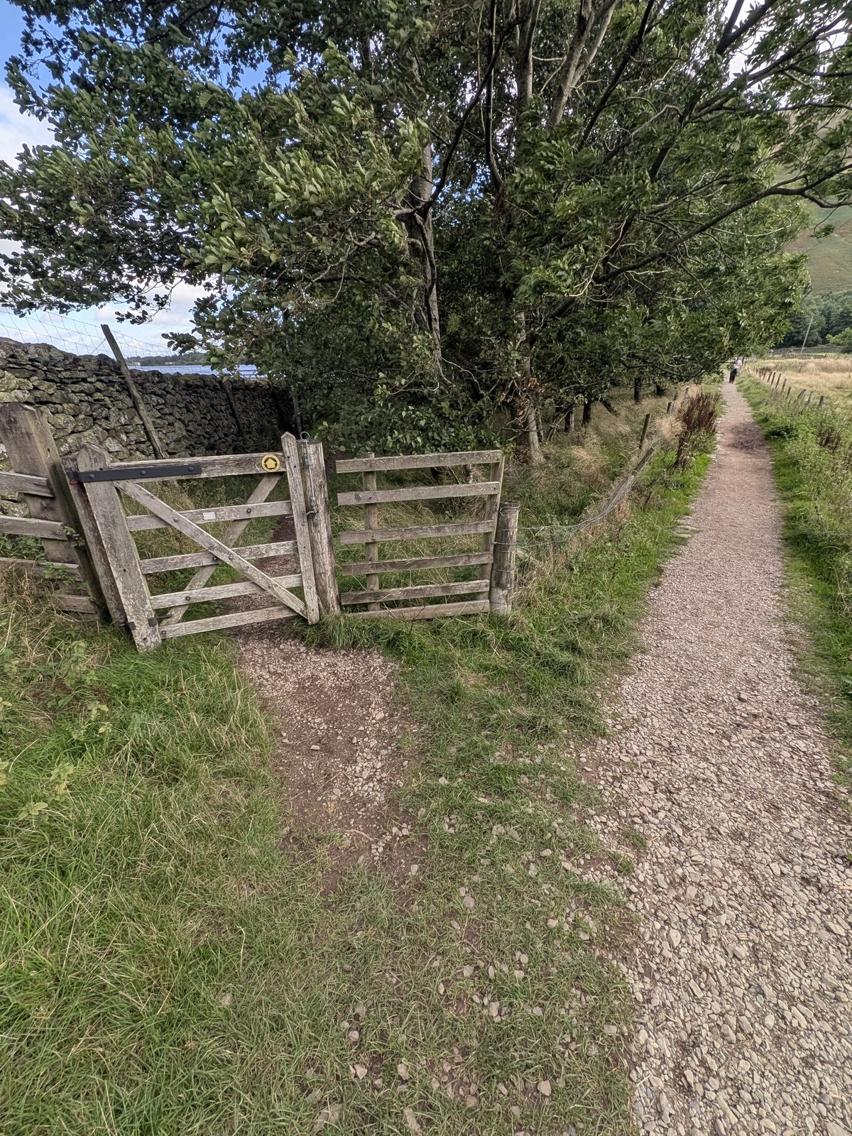 An open wooden five-bar gate set in a stone wall with a yellow waymark disc on the post, the main gravel path continuing to the right of it