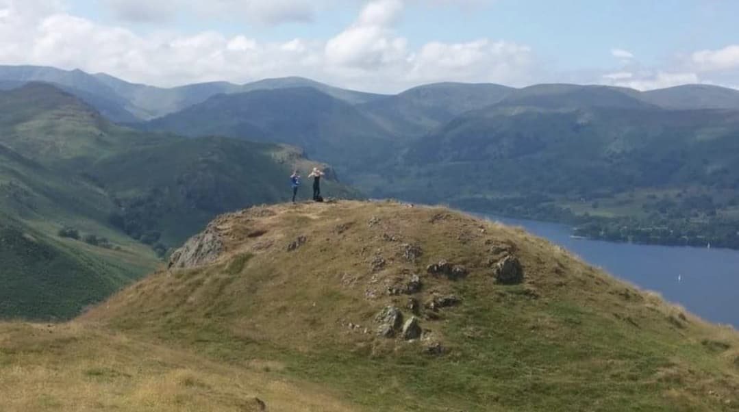 Walkers on the grassy summit of Hallin Fell with Ullswater and the fells beyond