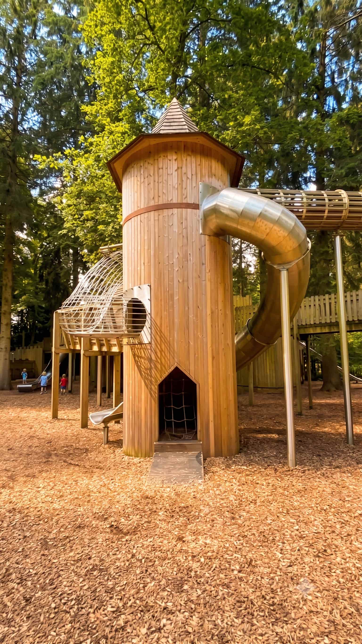 A timber turret at the Lost Castle adventure playground at Lowther, with a shingled conical roof, a stainless steel tube slide and a caged walkway running off into the trees