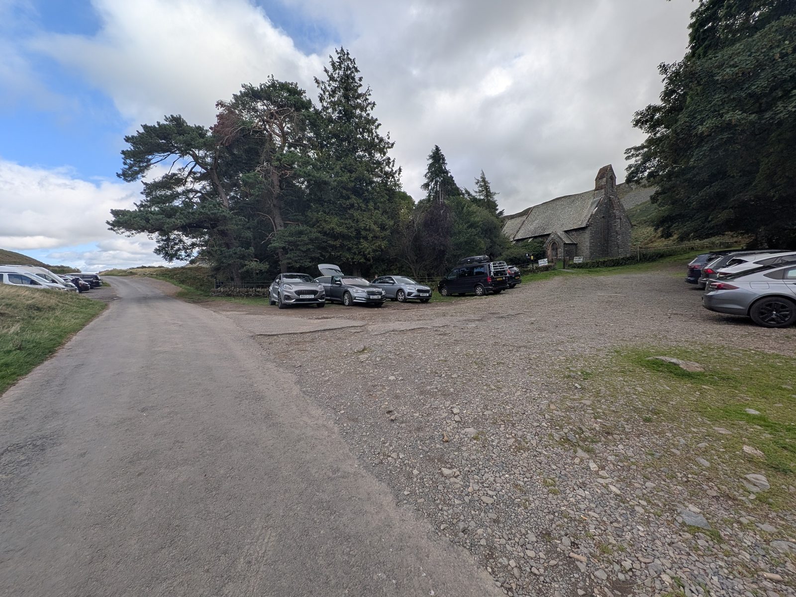 Gravel parking areas either side of the road at the top of the Hause, cars parked on both sides, St Peter's Church to the right