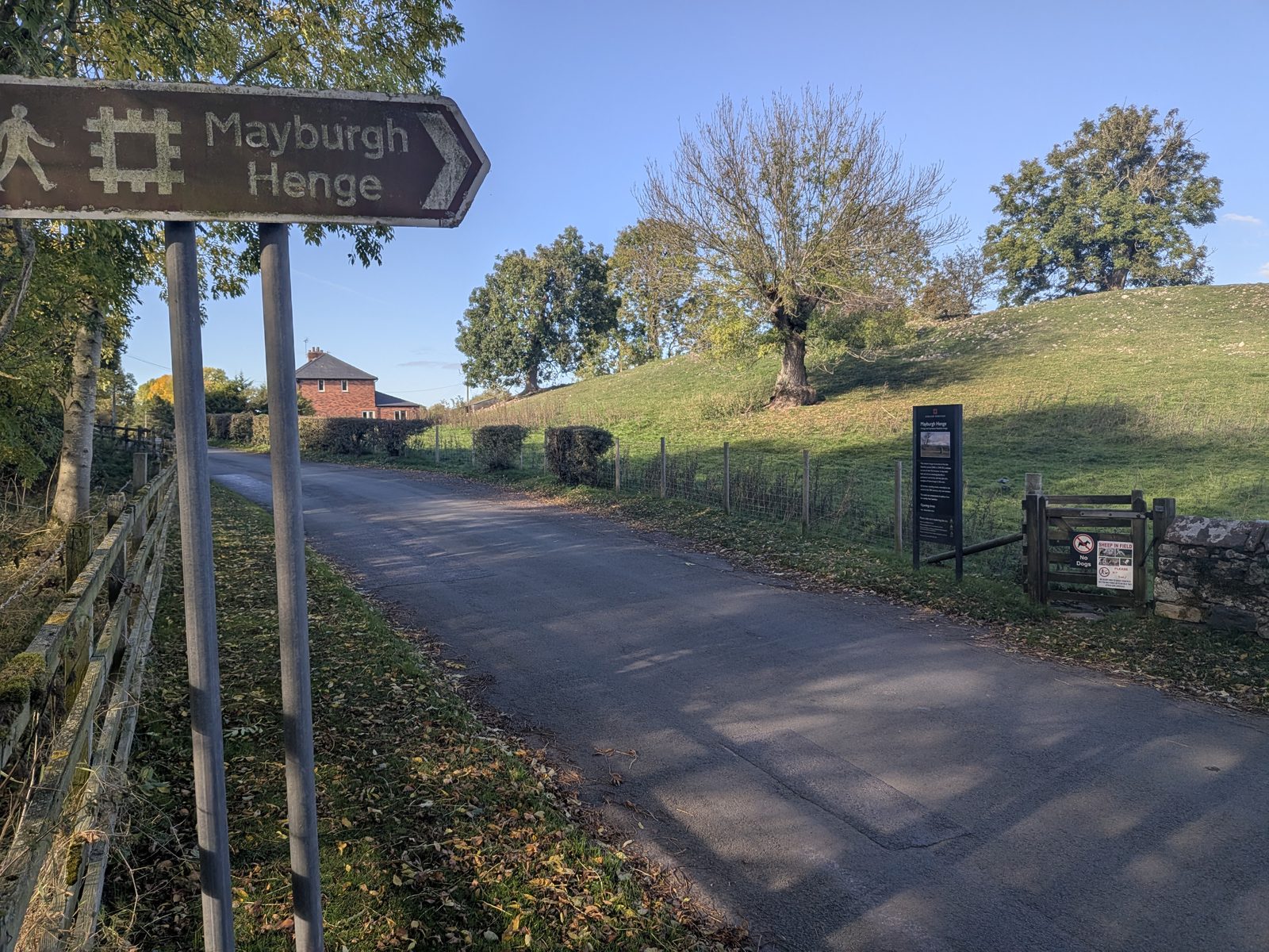 A brown English Heritage fingerpost for Mayburgh Henge beside a lane, with the grassy henge bank and a gate ahead