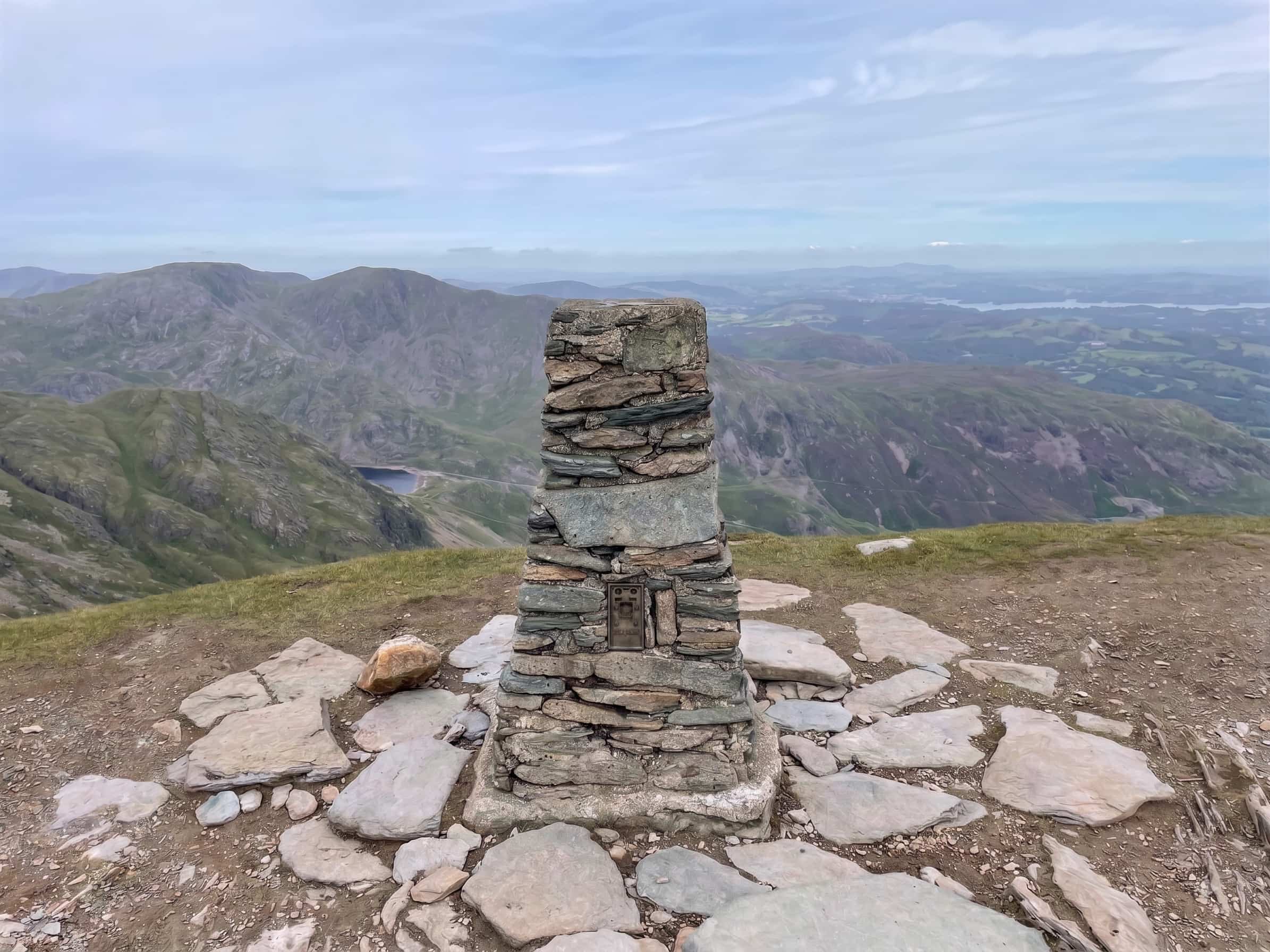 The stone-built summit column on the flagged top of the Old Man of Coniston, with Coniston Water and the fells stretching away beyond