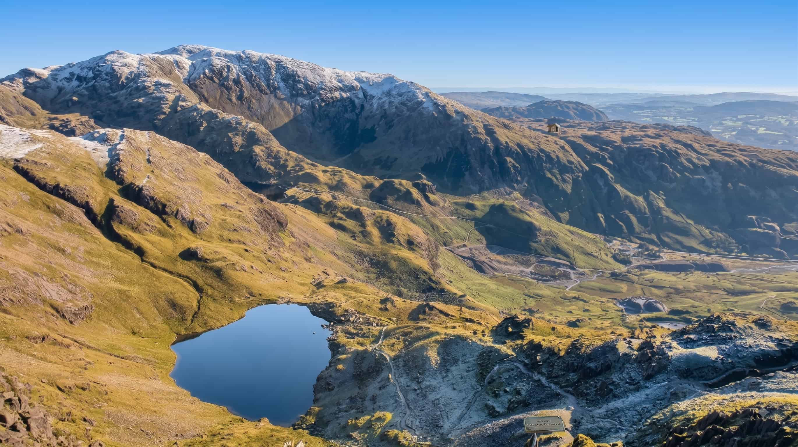A hanging tarn in its rocky bowl high on the Old Man of Coniston, with a dusting of snow on the ridge above and the quarry workings and valley below