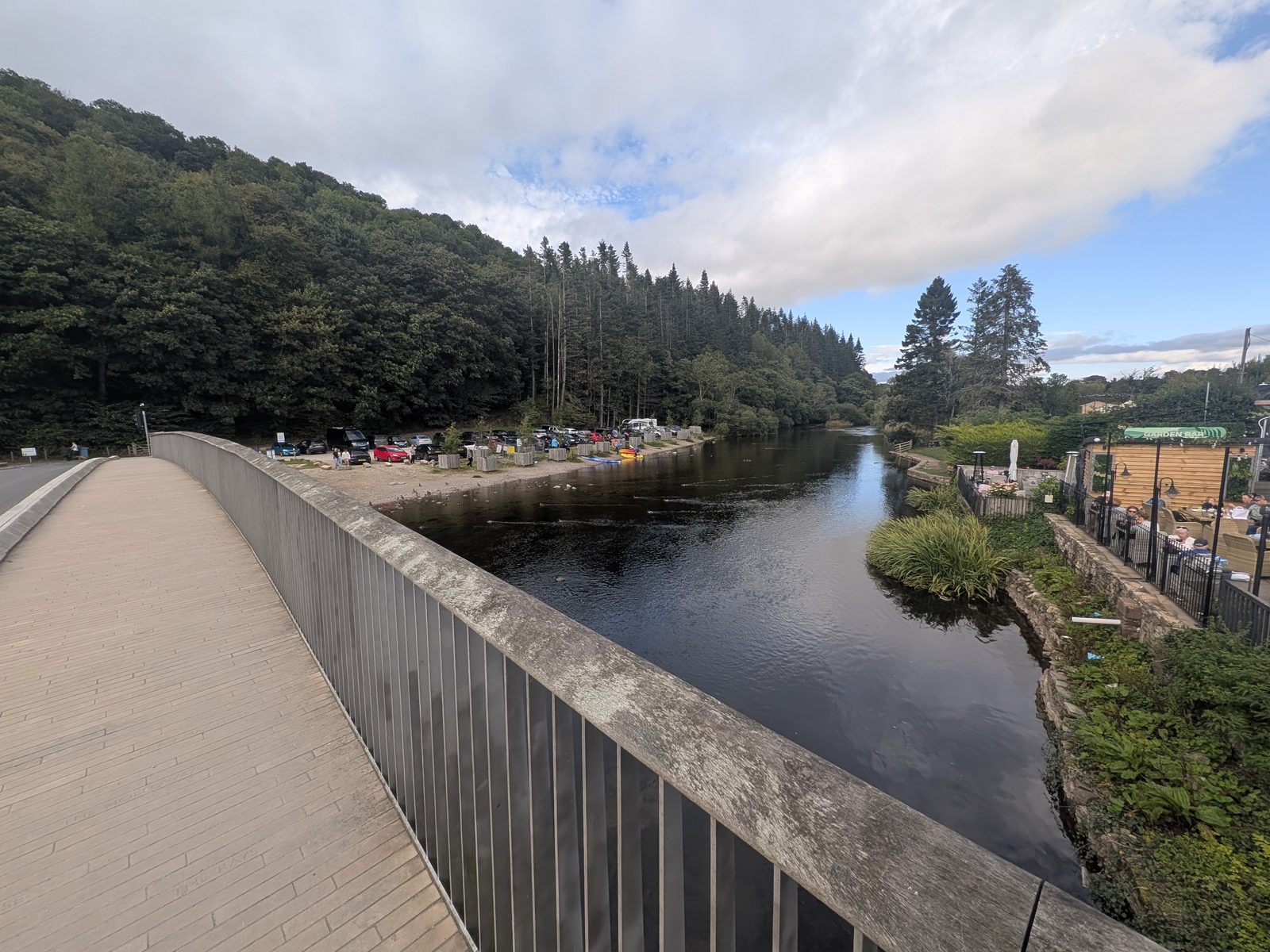 Looking down the River Eamont from the deck of the new Pooley Bridge, past the steel railing, towards Penrith — the wooded bank and moored boats on the left, away from the lake