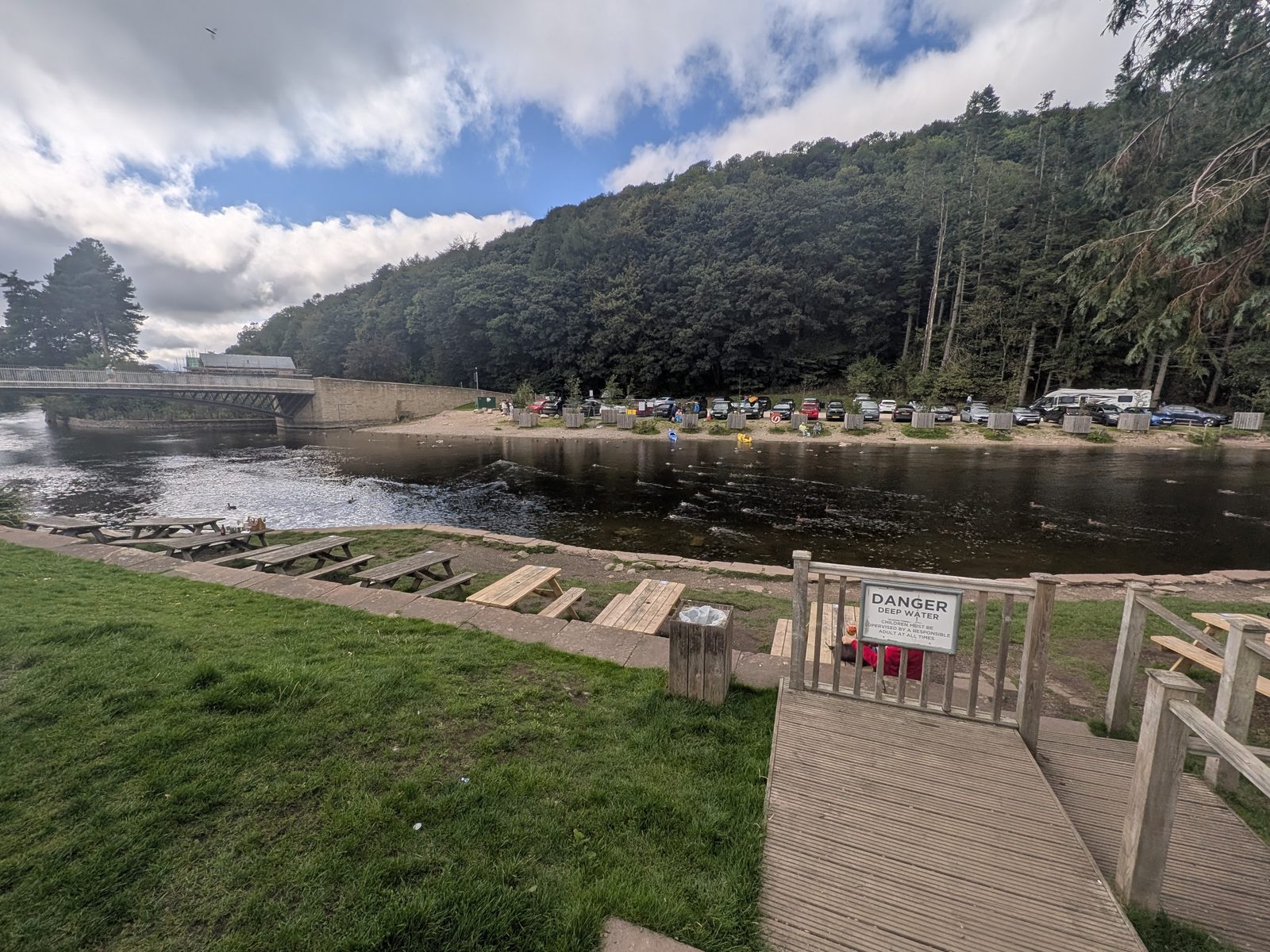 Picnic benches on the grass above the River Eamont at the Crown Inn, Pooley Bridge, with a Danger Deep Water sign, the road bridge and the wooded bank opposite
