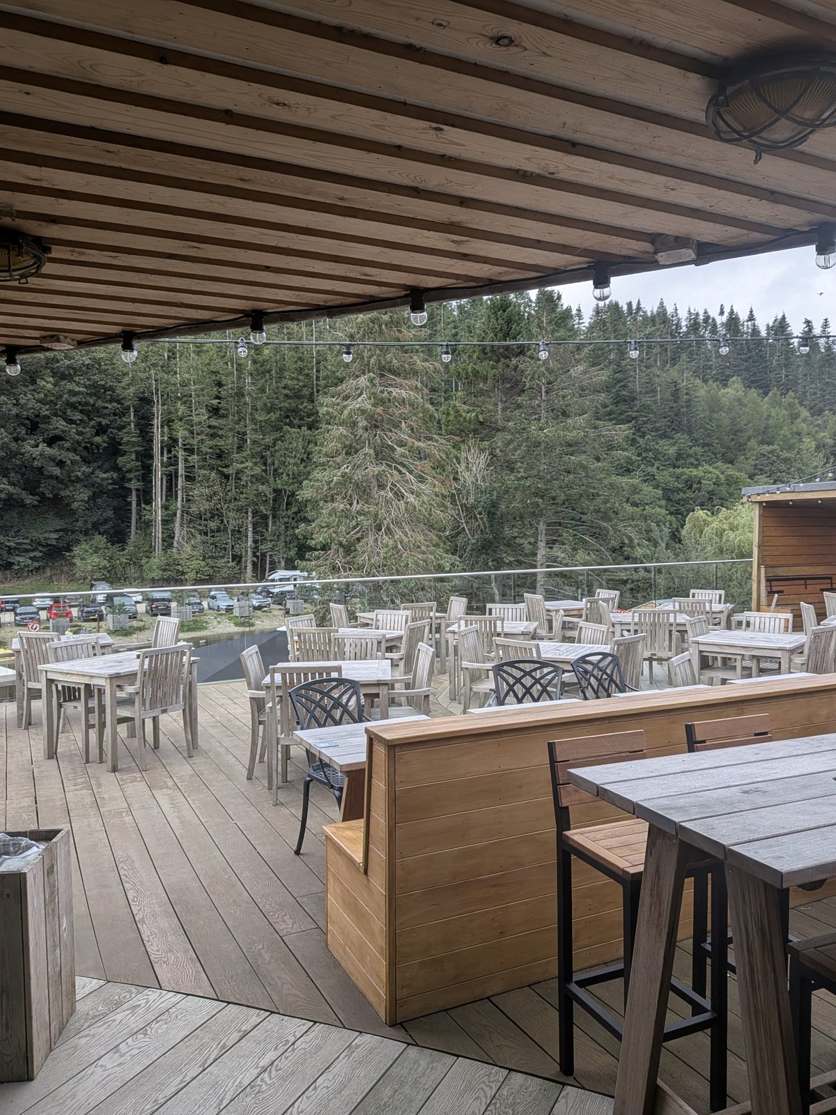 The covered timber terrace at the Crown Inn, Pooley Bridge, with rows of wooden tables and chairs under festoon lights and a glass balustrade looking out to the trees