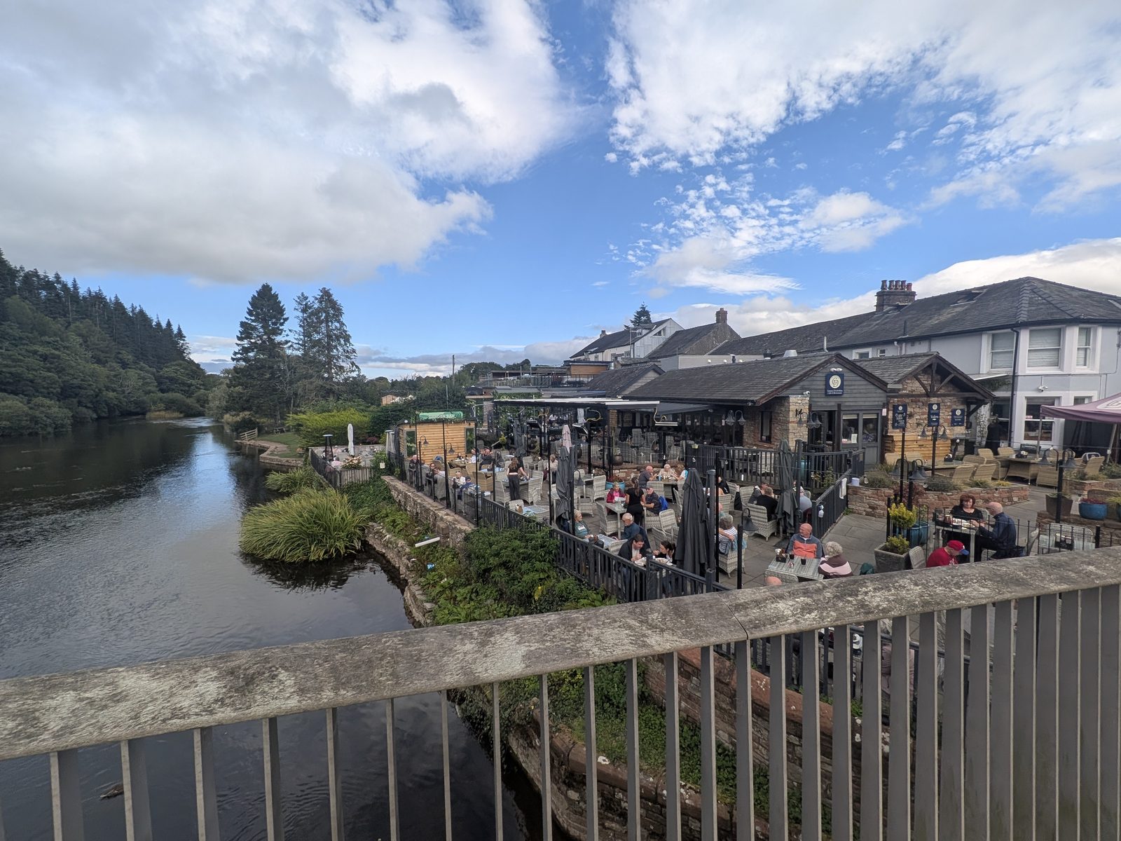 Riverside terrace tables full of diners under umbrellas on the bank of the River Eamont at Pooley Bridge, seen from the bridge, with the wooded far bank and the lake beyond