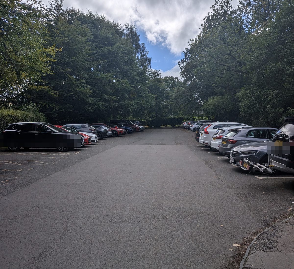 The main village car park at Pooley Bridge, a tarmac area lined with parked cars under mature trees