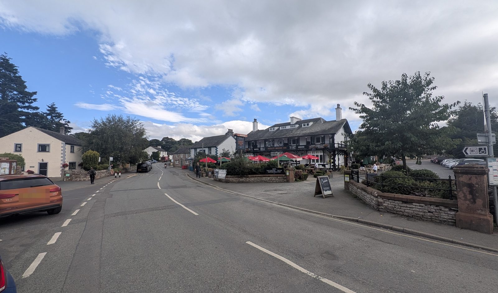 The main street through Pooley Bridge village, with a whitewashed cottage on the left, an inn with red umbrellas on its forecourt ahead and trees behind