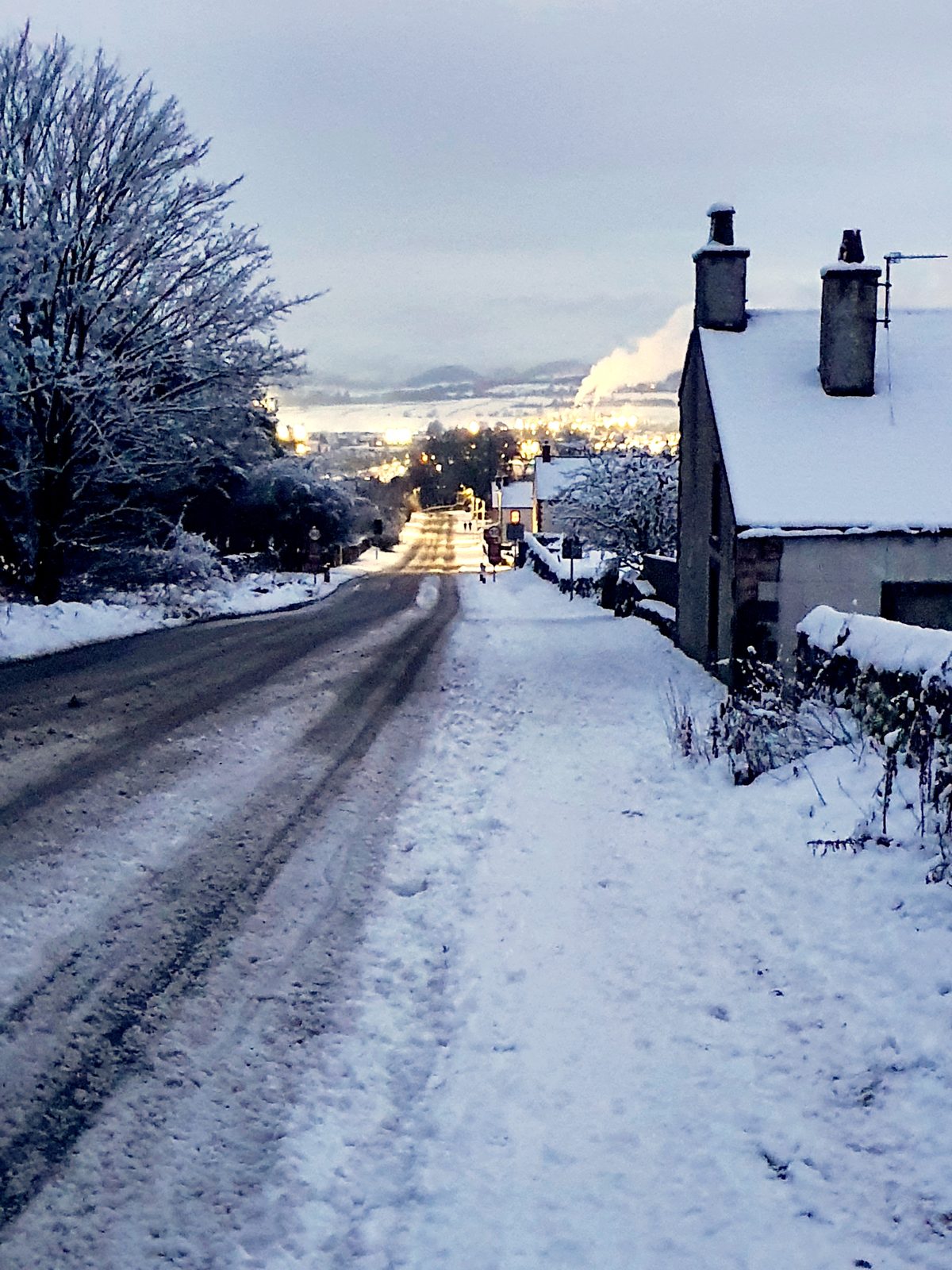 Salkeld Road in snow at dusk, with the lights of Penrith below and chimney smoke rising