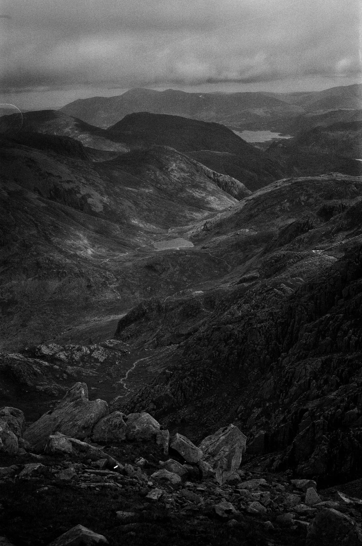 Black and white view from high on Scafell Pike across rugged fells, with a small tarn in the valley below and a lake in the far distance