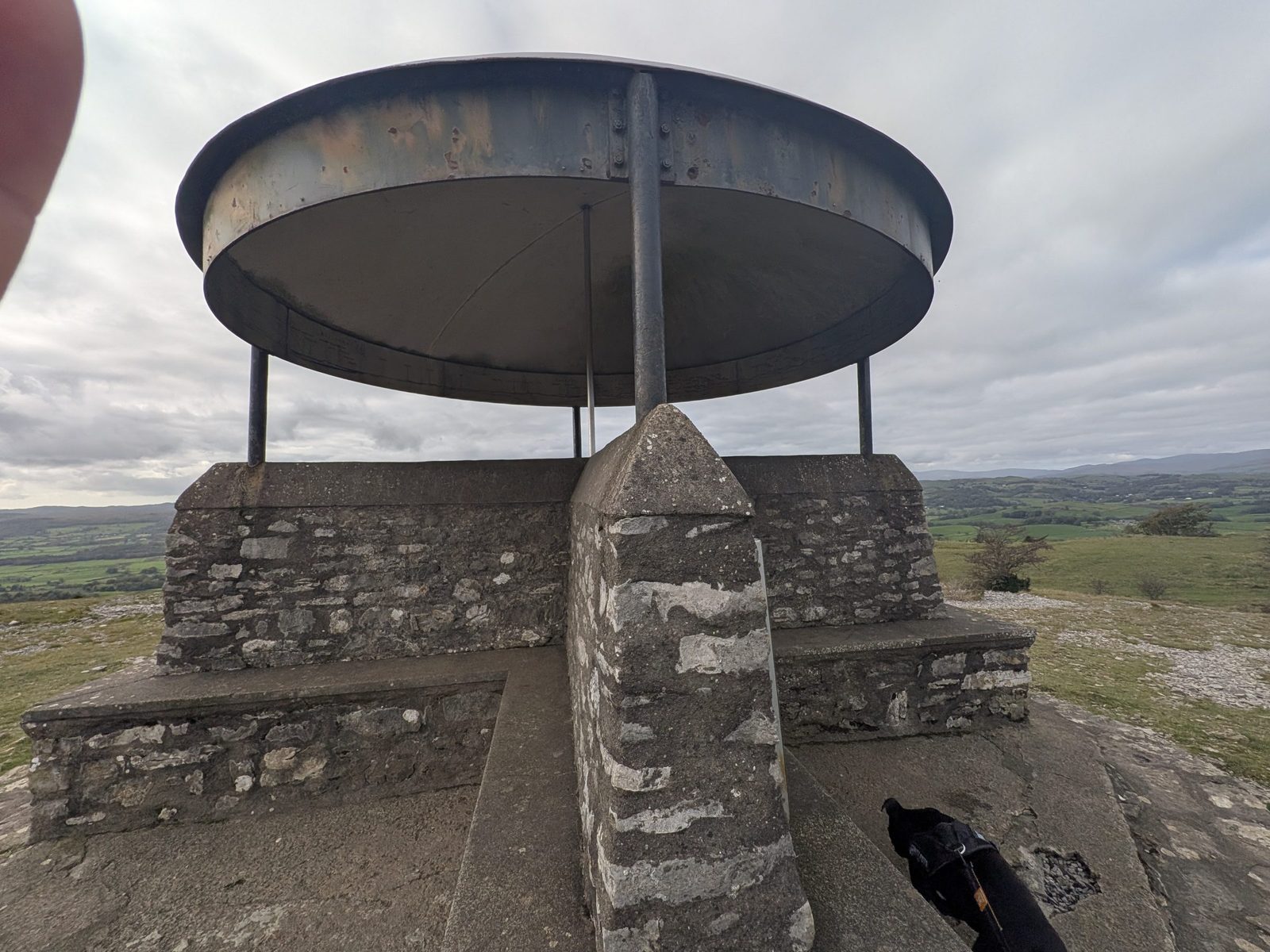 The Mushroom shelter on Scout Scar close up, with its stone base and metal roof, and Cass, a black Staffordshire Bull Terrier, in the corner