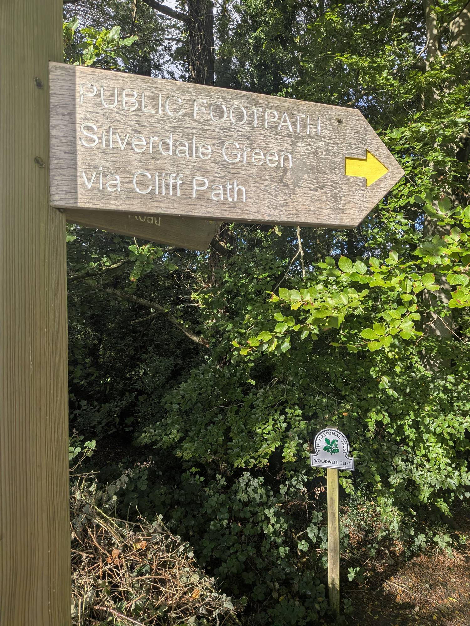 Wooden fingerpost reading Public Footpath, Silverdale Green via Cliff Path, with a National Trust Woodwell Cliff marker behind it