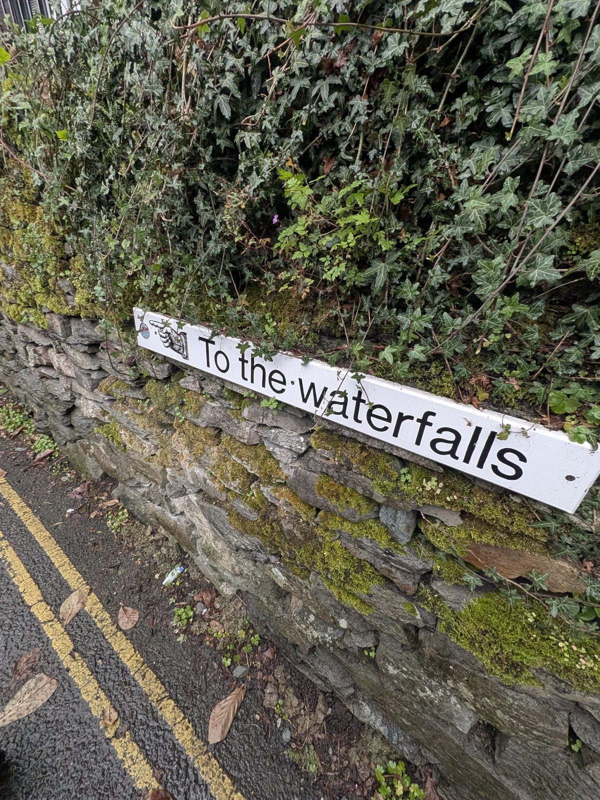 A white &ldquo;To the waterfalls&rdquo; sign fixed to a mossy slate wall beside double yellow lines
