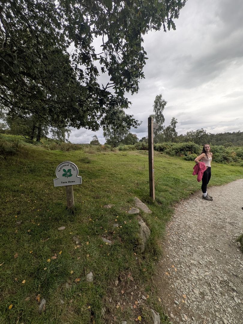 National Trust oak-leaf sign reading Tarn Hows beside a wooden fingerpost at the start of the surfaced path