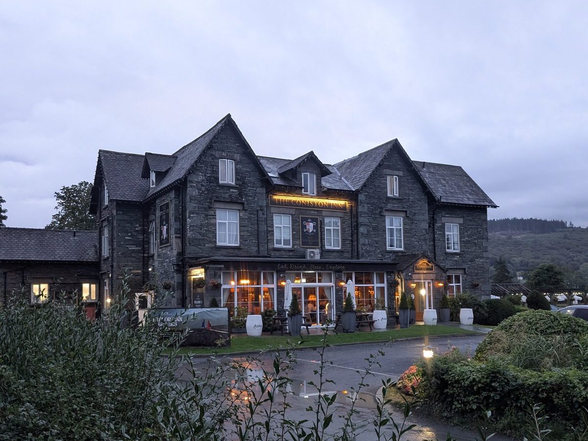 A large slate-built inn in Coniston with its ground-floor windows lit at dusk, planting in the foreground