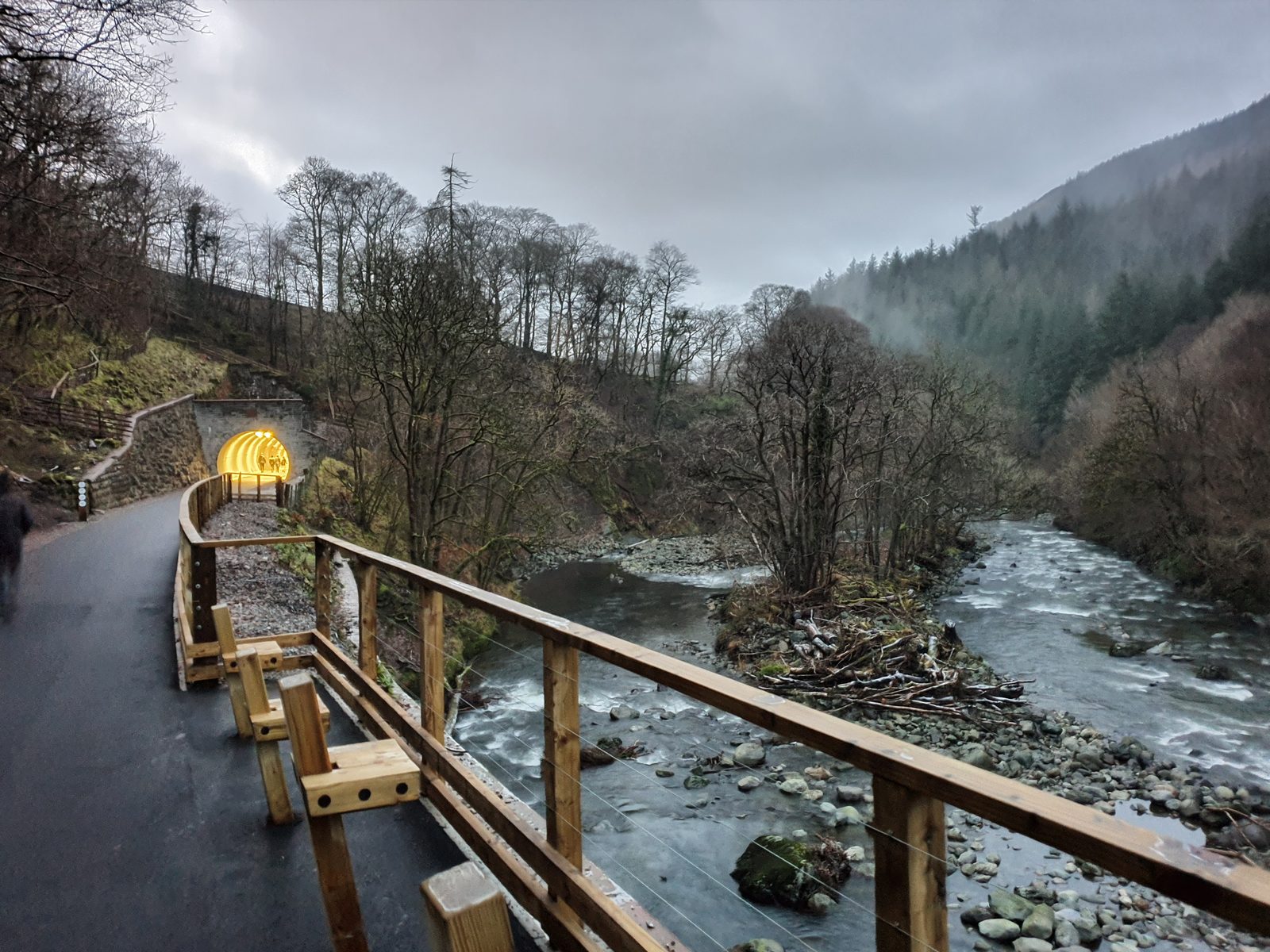 The Keswick to Threlkeld railway trail curving towards the lit tunnel mouth, with the River Greta running below and wooded fellside beyond