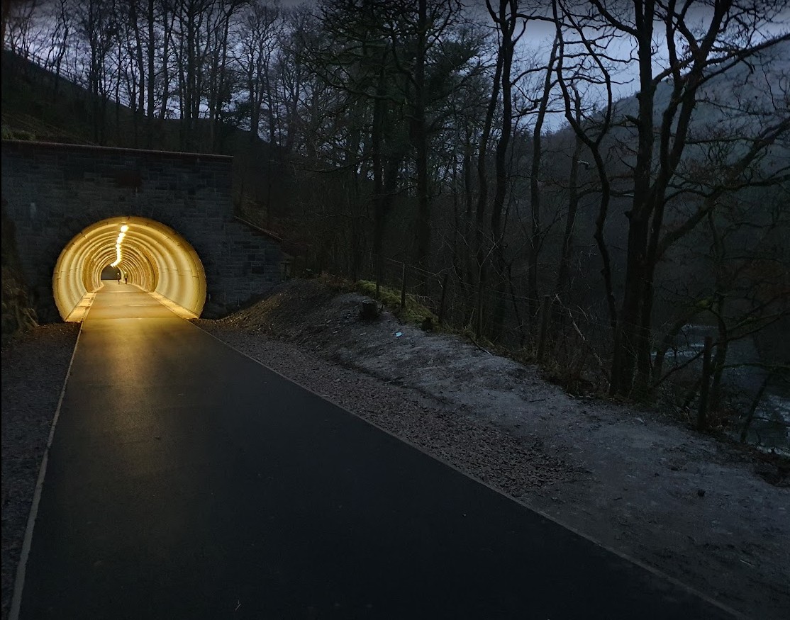 The lit tunnel mouth on the Keswick to Threlkeld railway trail at dusk, the surfaced path running in past a stone portal with bare winter trees above
