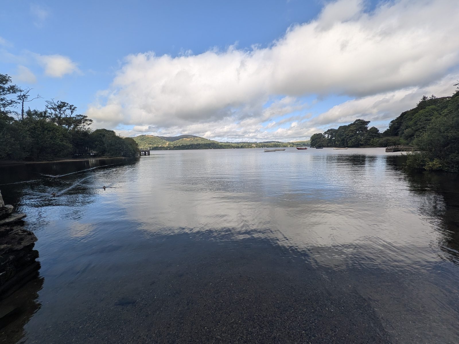 A shingle shore at water level on Ullswater, the water flat calm, with jetties and moored boats in the distance