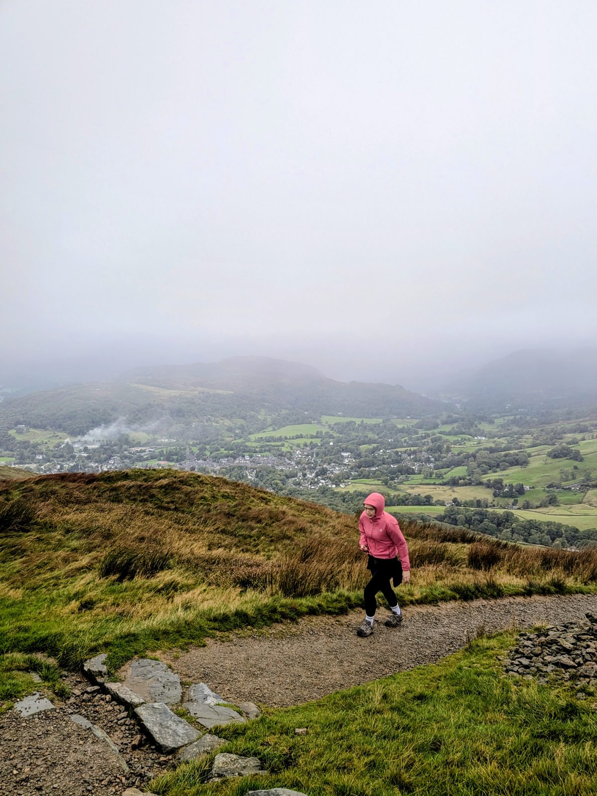Ellie in a pink jacket walking along the path, with Ambleside and the valley below in mist