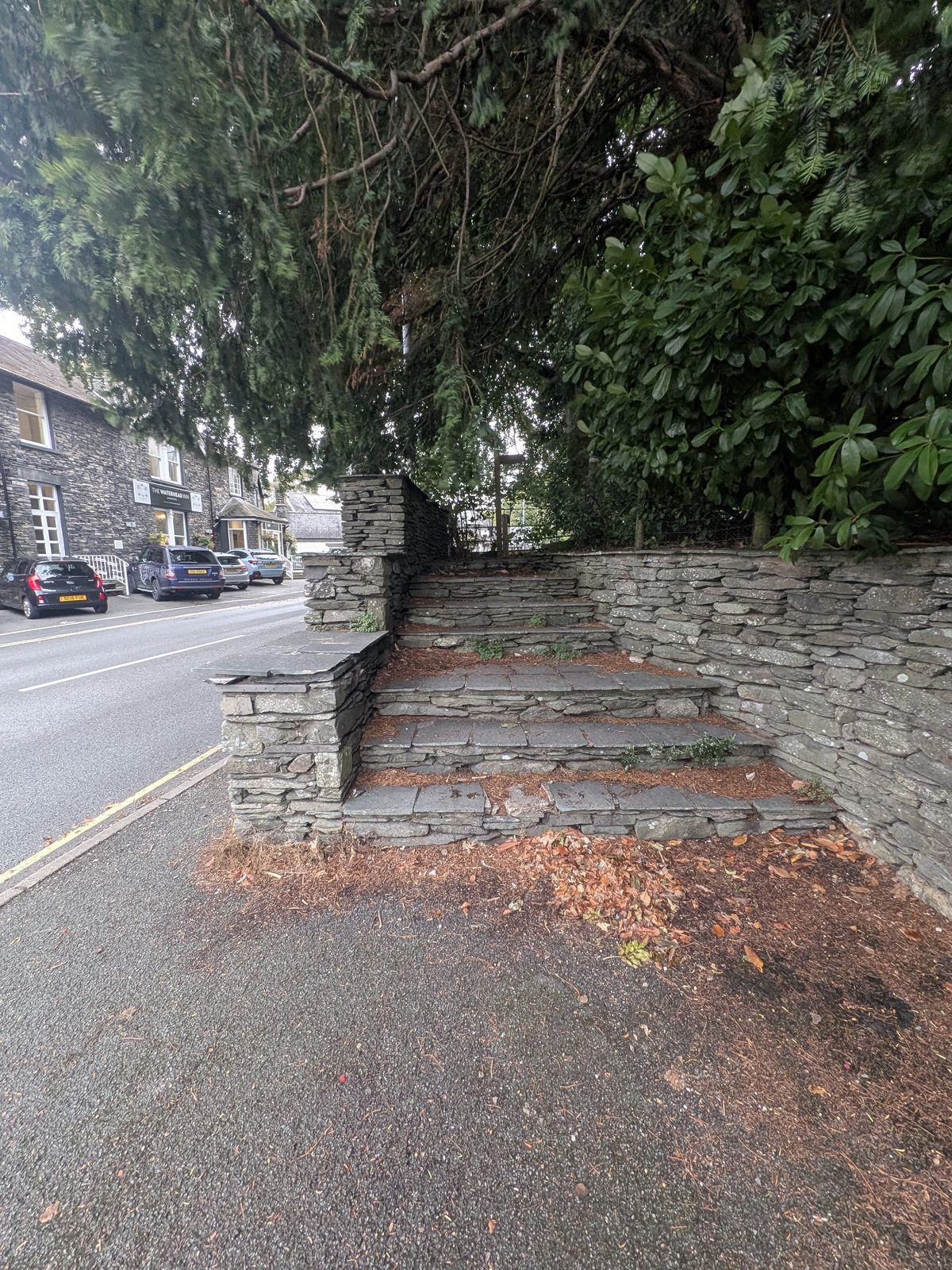 Stone steps rising from the pavement of the A591 beside a stone wall and trees