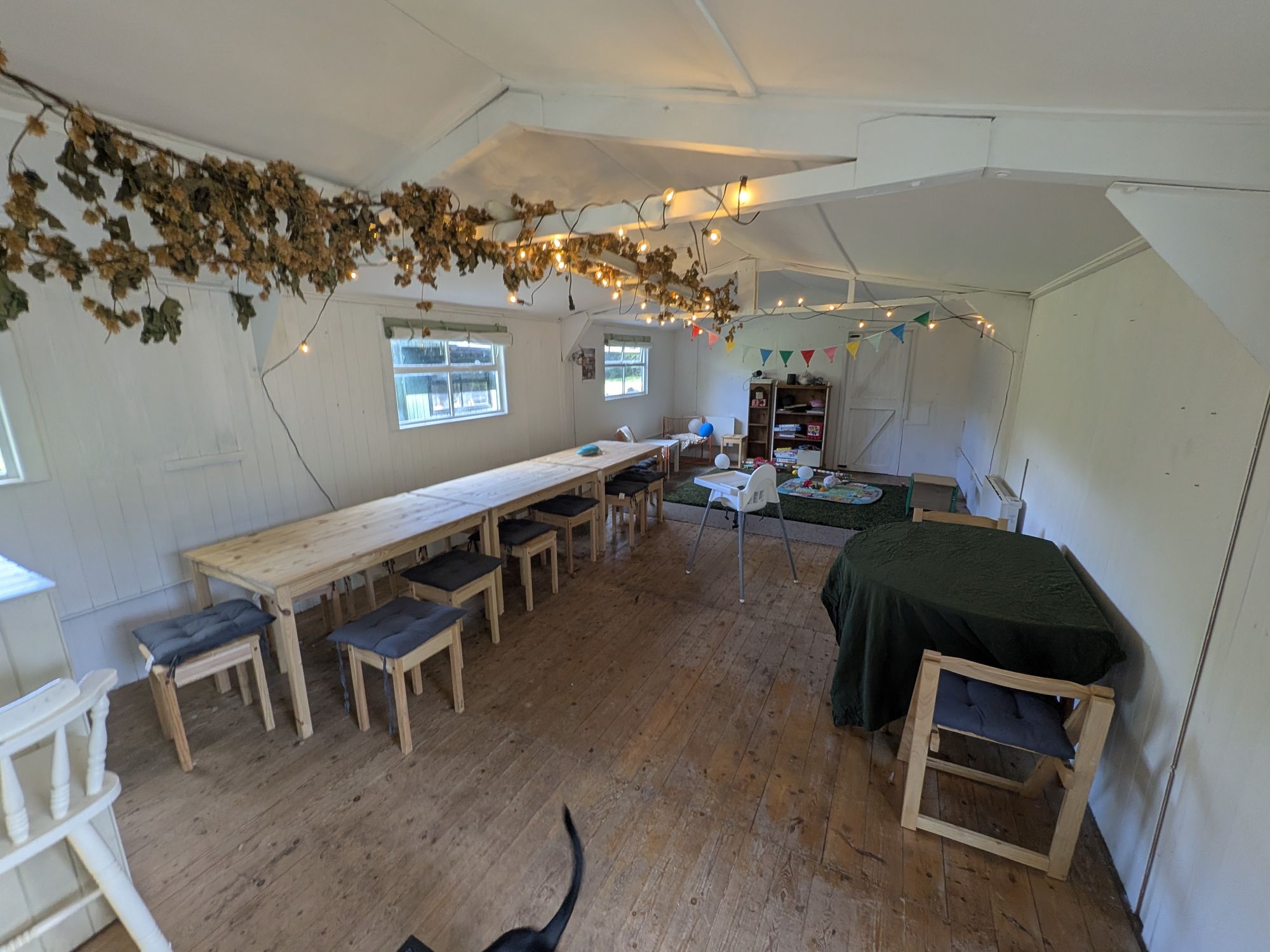 Long pine tables and stools inside The Bau garden room at The Wolf Cafe, with fairy lights and dried hops along the rafters