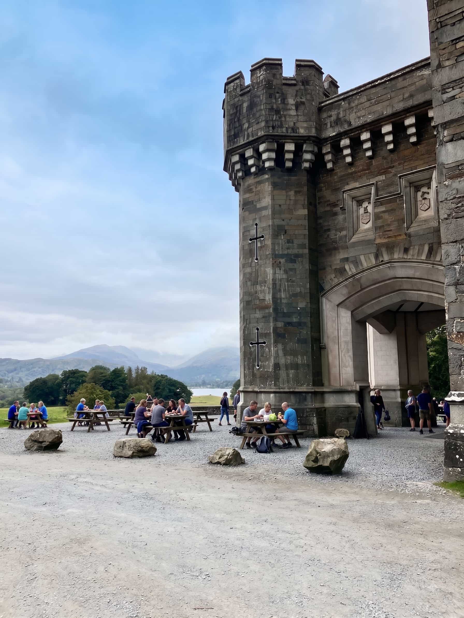 Picnic tables outside Joey's Café beneath the gatehouse arch at Wray Castle, with Windermere and the fells beyond